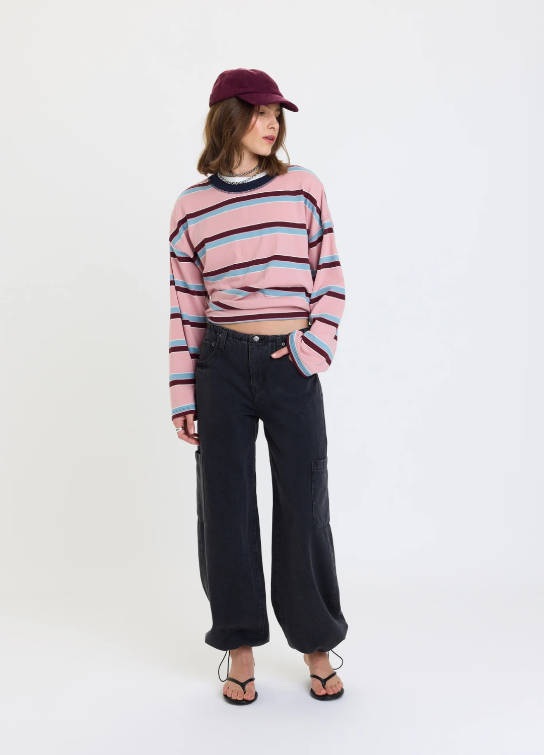 A model stands against a plain white studio background wearing a pink striped long-sleeve crop top, dark wide-leg pants, black sandals, and a burgundy cap.
