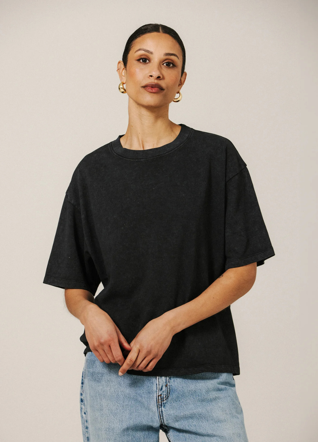 A woman wearing an oversized black crewneck t-shirt and light blue jeans, standing against a plain light beige studio background.